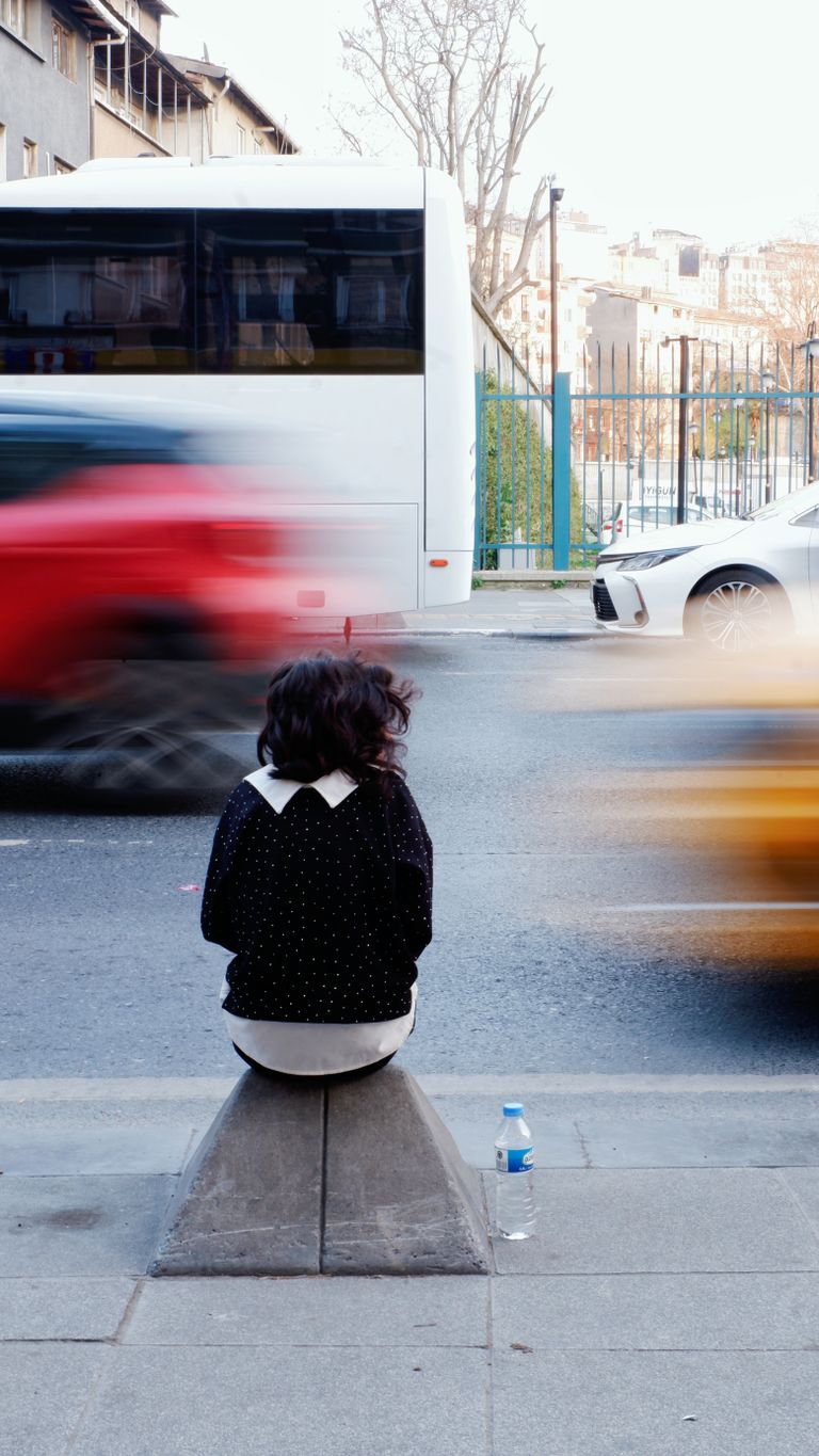 A person sits on a curbside watching traffic in a city street, with motion blur of cars.