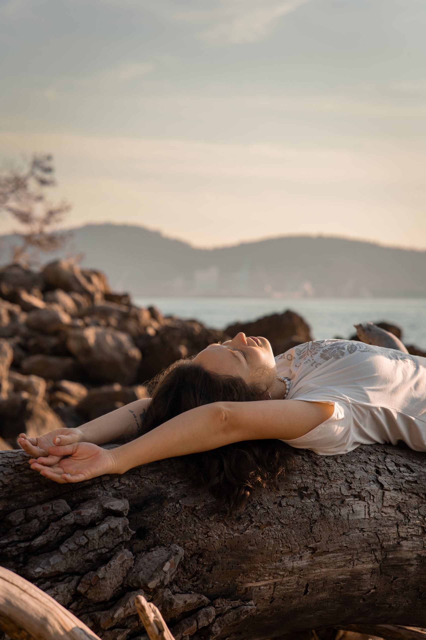 A woman lies on driftwood by the sea, embracing a peaceful sunset moment.