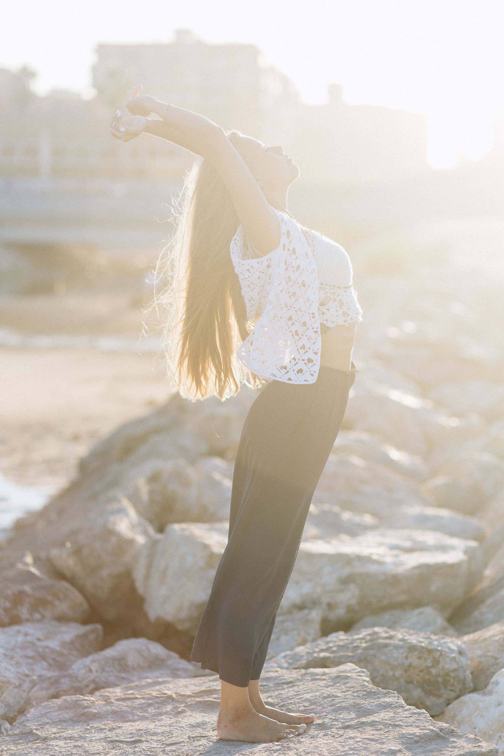 Young woman in peaceful meditation pose on a sunlit rocky shoreline, embracing tranquility and nature.