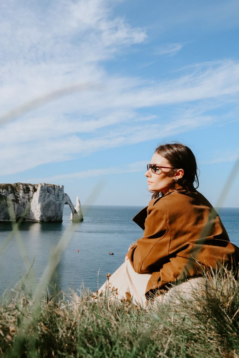 A woman in sunglasses gazes at the famous cliffs of Étretat in France. The scene is serene and picturesque.