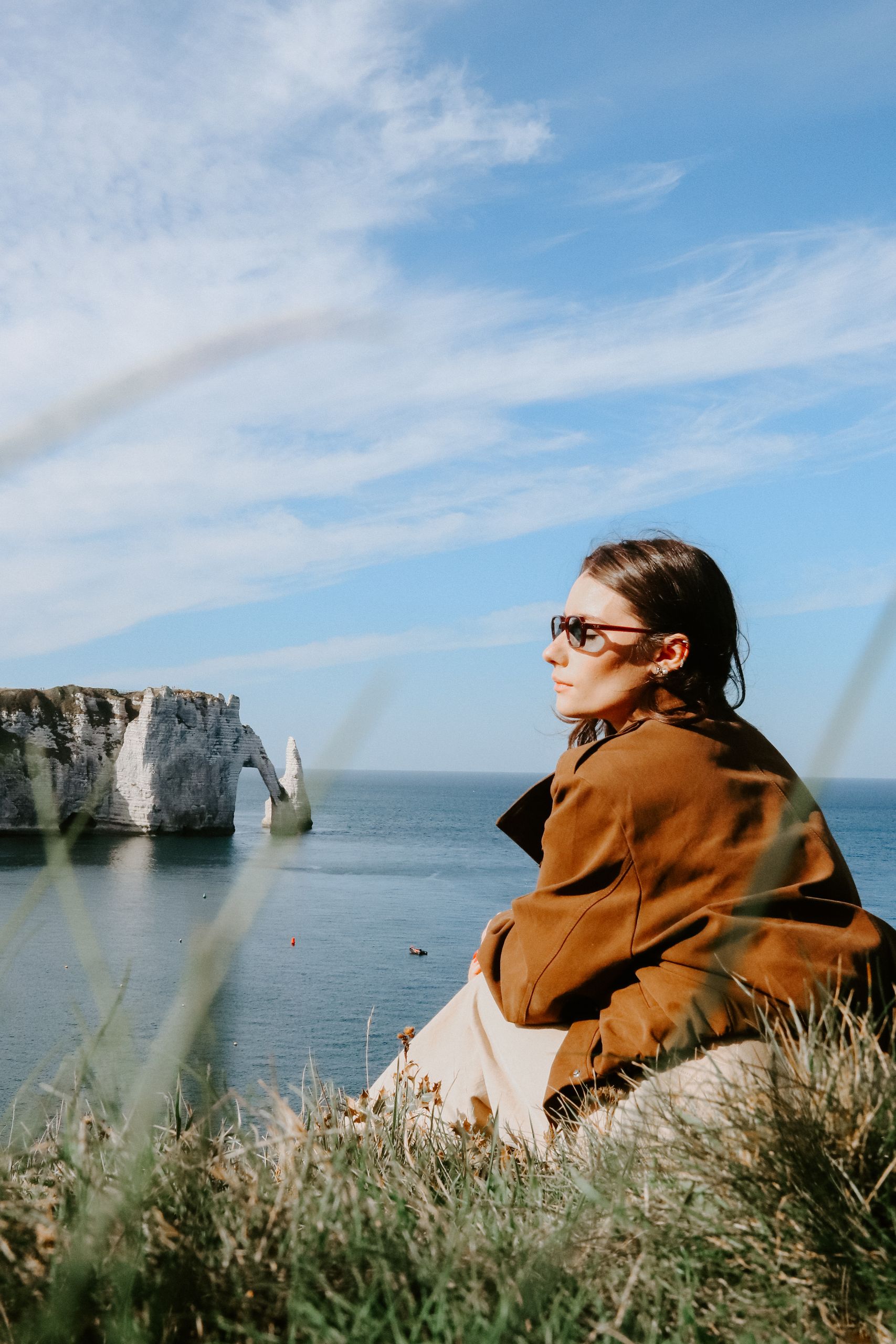 A woman in sunglasses gazes at the famous cliffs of Étretat in France. The scene is serene and picturesque.