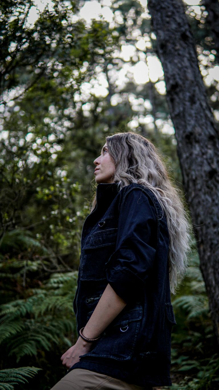 Side view of a woman with long blonde hair walking in a serene forest, sporting a denim jacket.