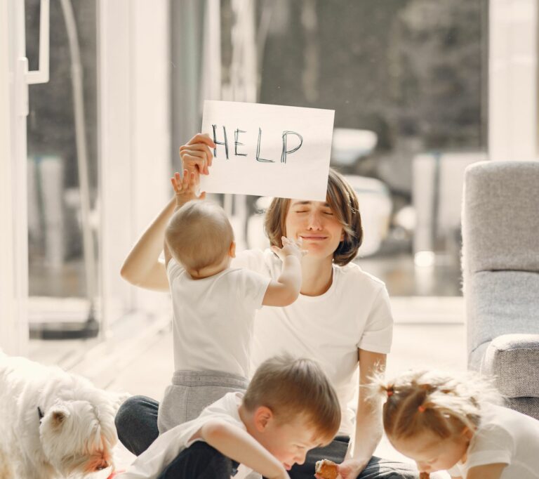 Woman holding ironic placard asking for help above head while sitting on floor with children playing in well lit living room