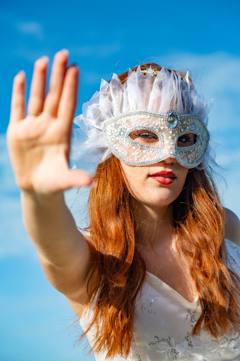 Woman in an elegant masquerade mask extending hand outdoors with a bright sky backdrop.